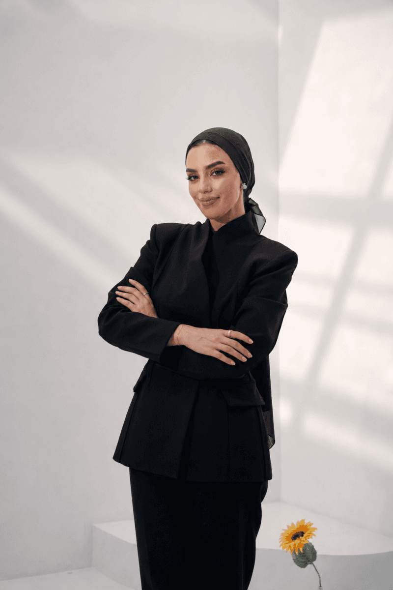 ISHRAQ BRAHMI in a black tailored suit, standing with folded arms in a sunlit studio